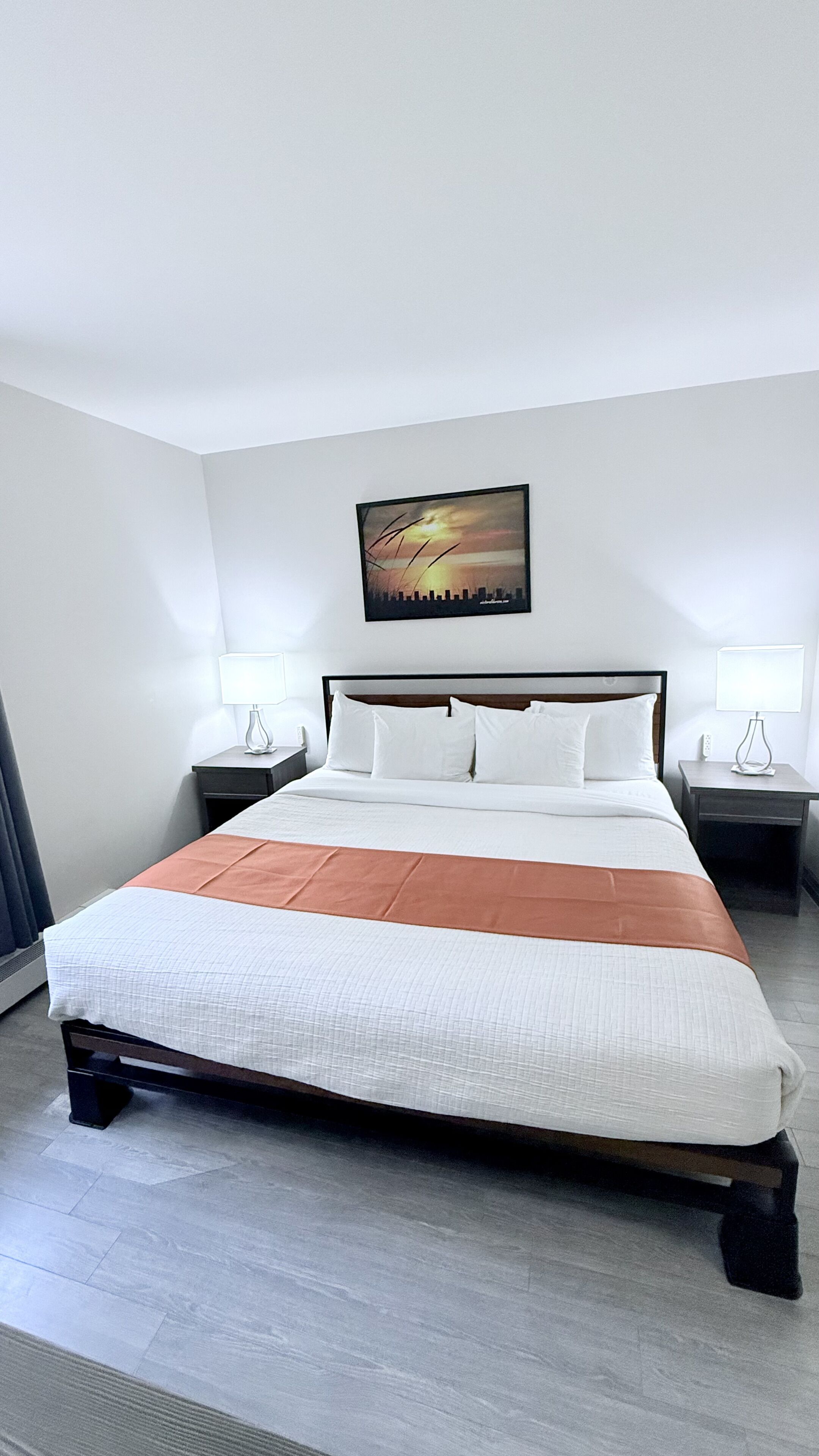 Una habitación de hotel con una cama grande, dos mesitas de noche con lámparas, un cuadro de paisaje urbano en la pared y un diseño moderno.