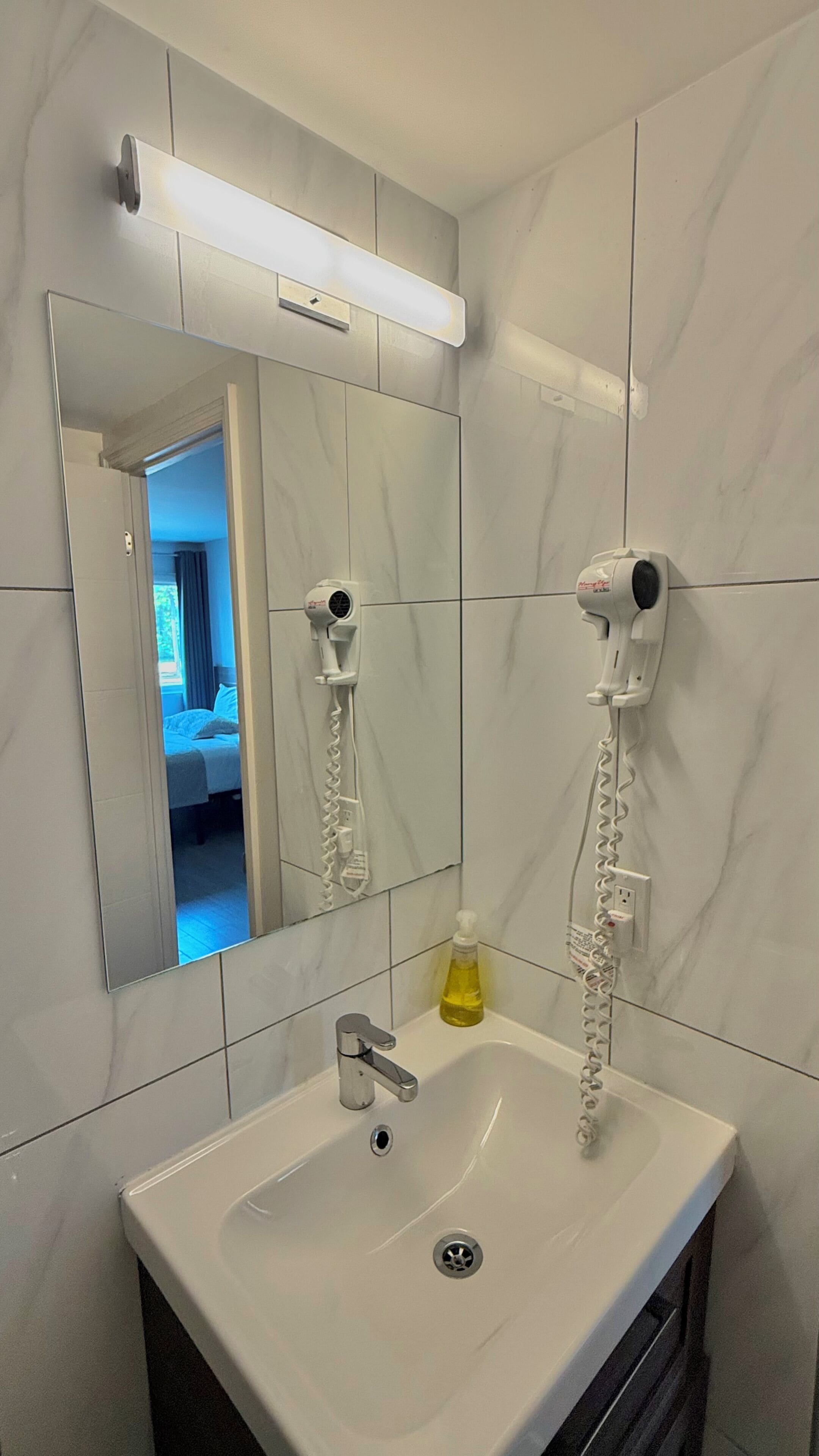 A bathroom with a white sink, a mirror, two hair dryers mounted on the wall, and a bottle of soap on the sink.