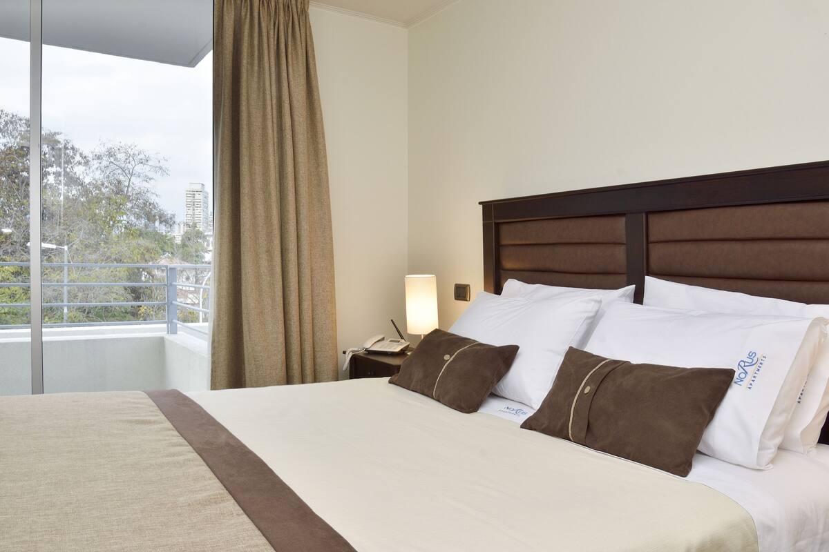 Habitación de hotel con una cama grande, una mesita de noche con lámpara y vistas al exterior a través de una ventana.