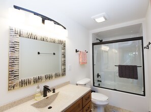 A bathroom with a large mirror, a sink with a soap dispenser, a toilet, and a glass-enclosed shower area.