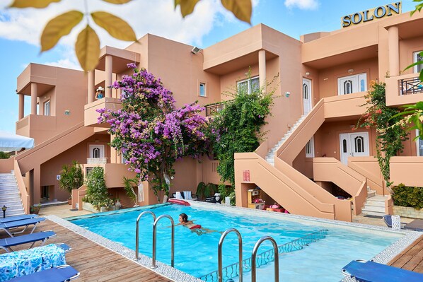 A swimming pool with a person swimming, surrounded by a building with a 'SOLON' sign, stairs, and a wooden deck with lounge chairs. - Solon Apartments (Platanias)