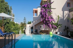 A swimming pool with a wooden deck, a covered seating area, and a building with purple flowering vines. - Solon Apartments (Platanias)
