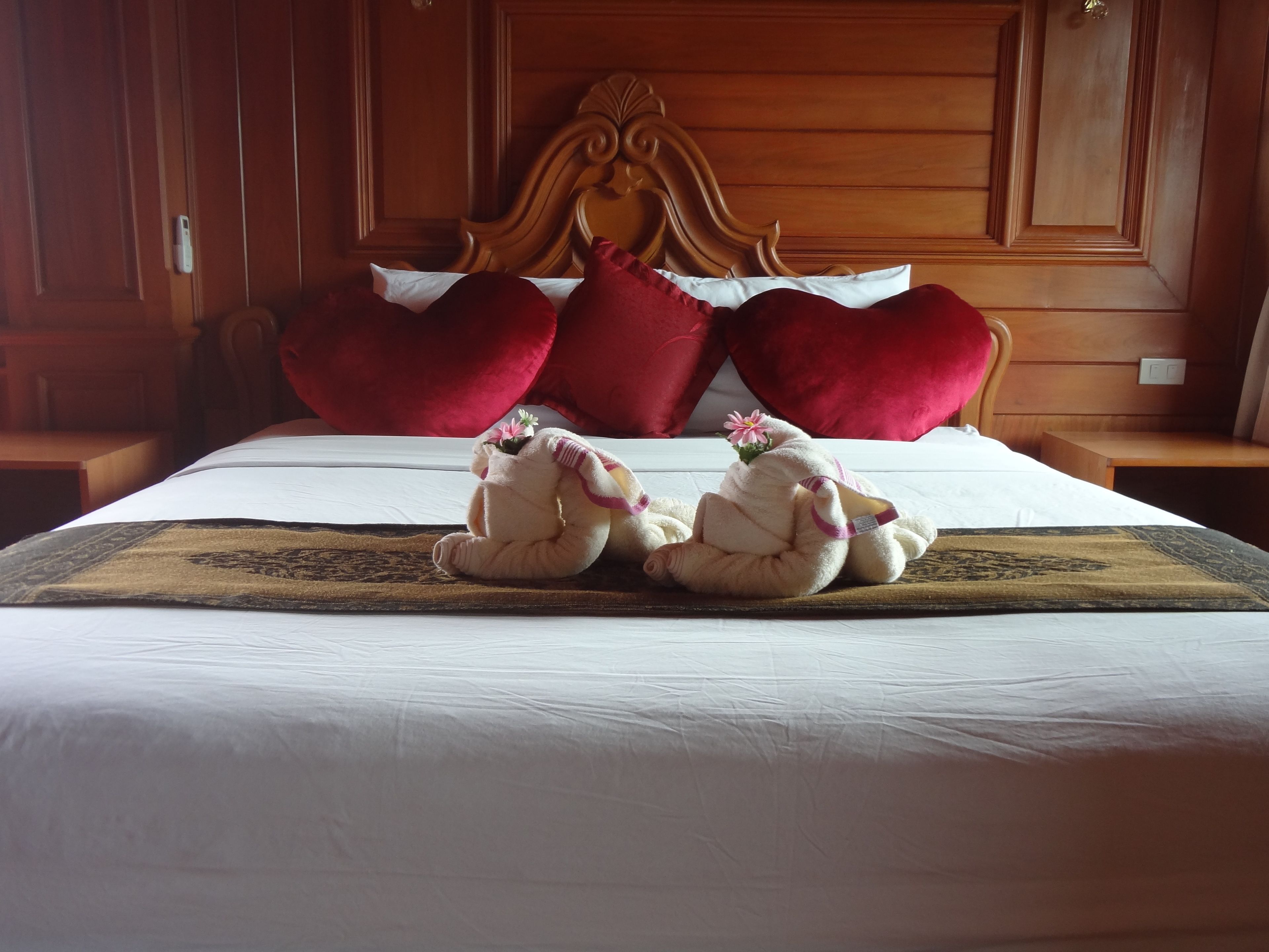 A neatly made bed with red pillows, a wooden headboard, and two towels shaped like elephants with flowers.