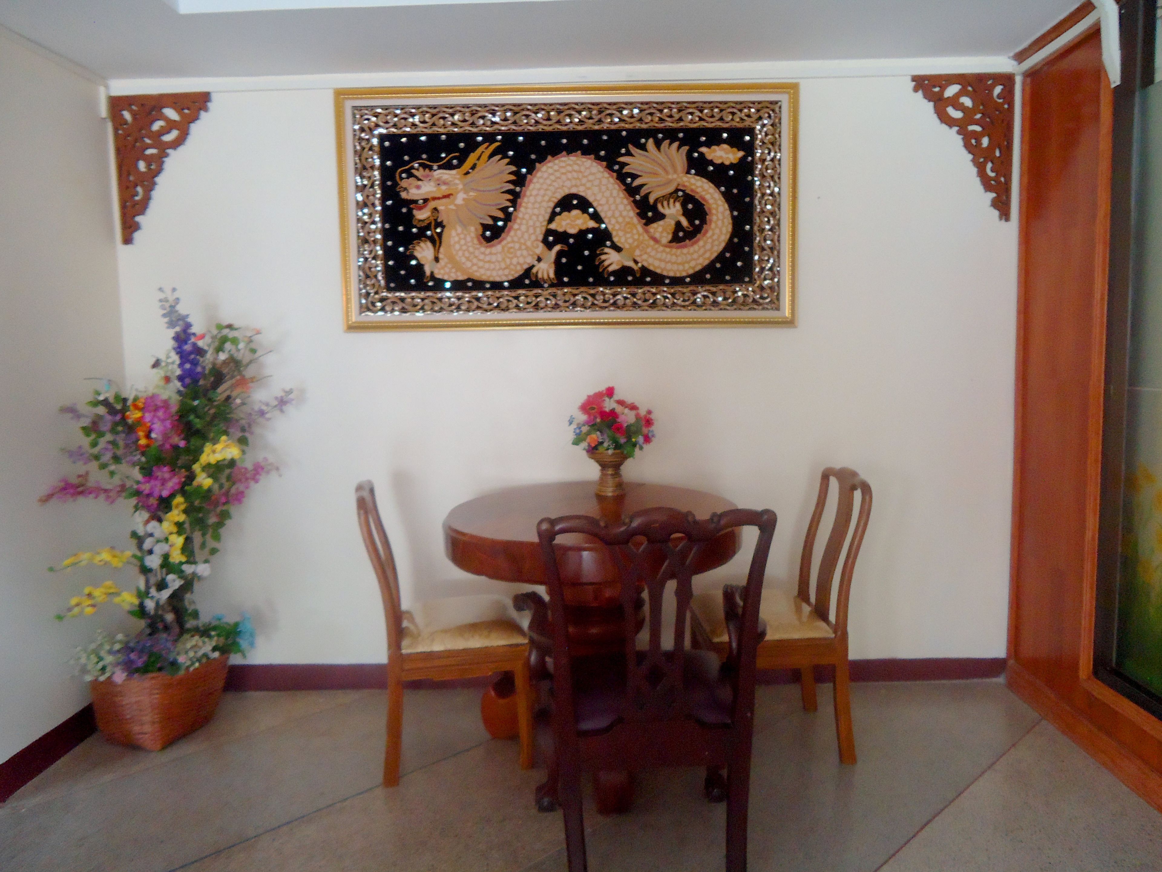 A dining area with a round wooden table, four chairs, a vase with flowers, and a framed dragon artwork on the wall.