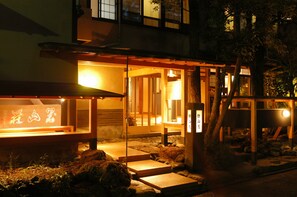 Edificio japonés tradicional con estructura de madera, interior iluminado y escalones de piedra que conducen a la entrada.
