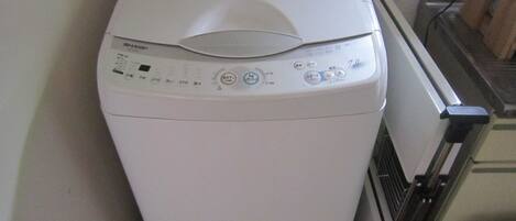 A white front-loading washing machine with control panel, located in a corner of a room with a radiator and a wooden cabinet.
