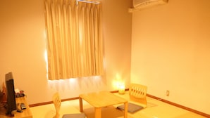 Una habitación con una mesa de madera, dos sillas, una lámpara pequeña, un televisor y una ventana con cortinas.
