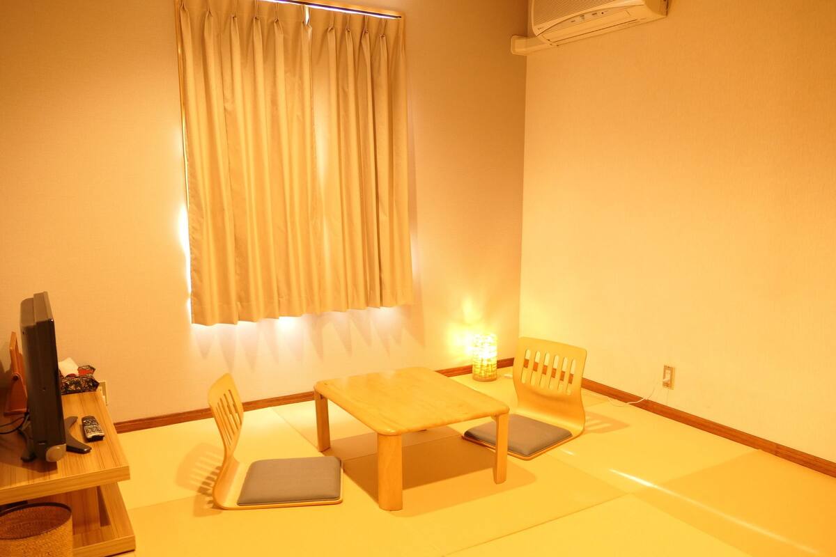 Ein Zimmer mit einem Holztisch, zwei Stühlen, einer kleinen Lampe, einem Fernseher und einem Fenster mit Vorhängen.