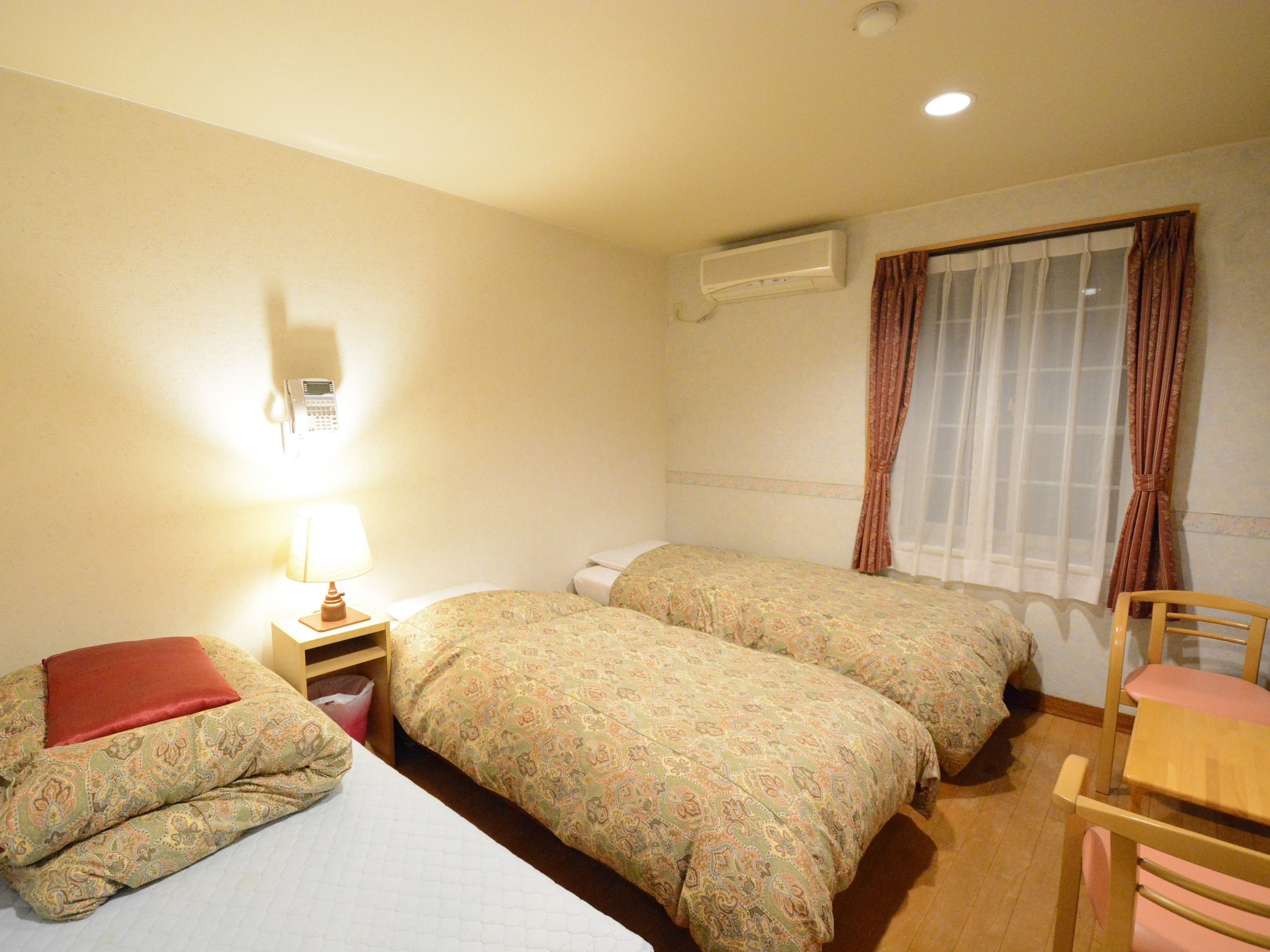 Une chambre d’hôtel avec deux lits, un bureau, une chaise, une lampe, un climatiseur et une fenêtre avec des rideaux.