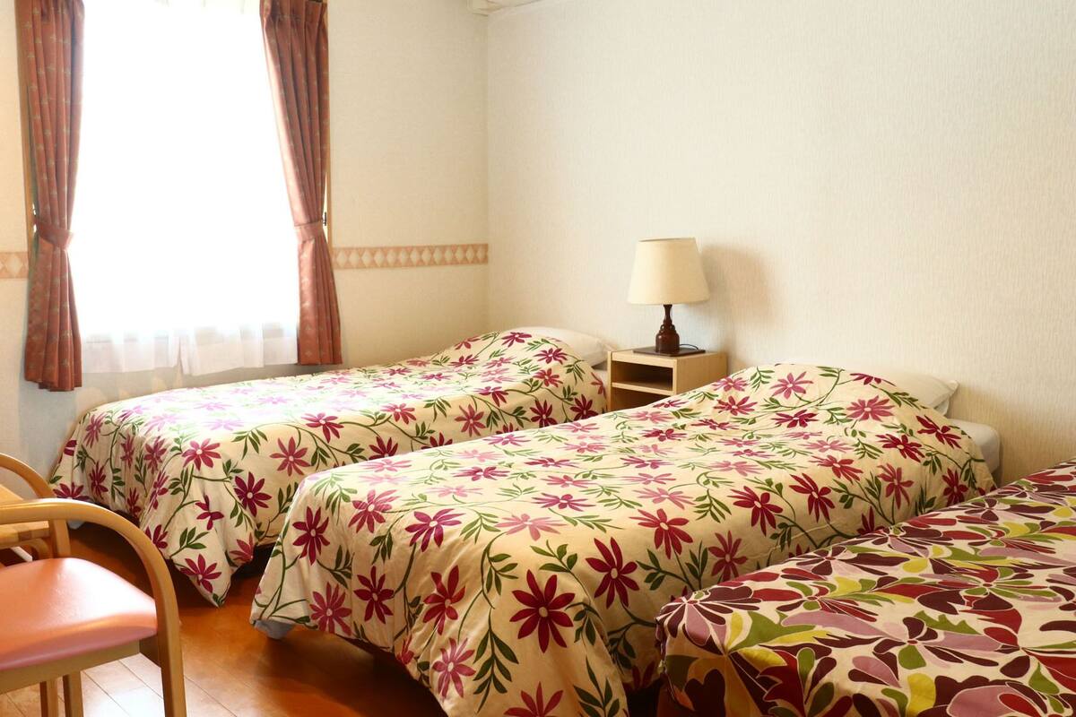 Ein Zimmer mit zwei Betten, einem Sessel, einem Nachttisch mit Lampe und einem fensterbepflanzten Fenster.