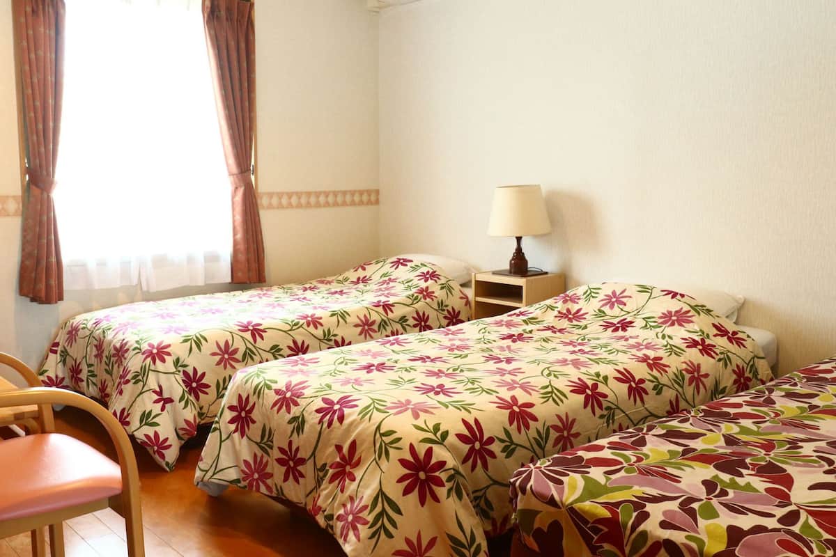 A room with two beds, a chair, a nightstand with a lamp, and a window with curtains.