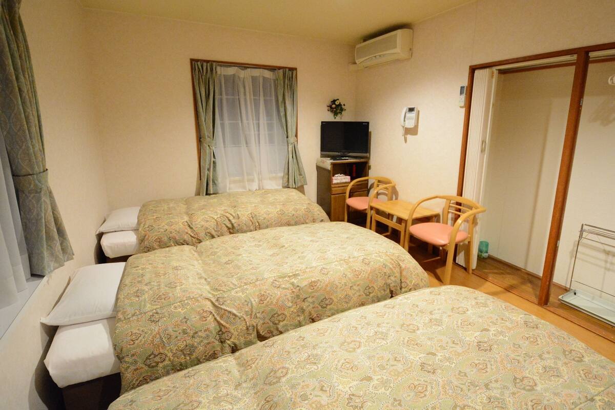 Ein Hotelzimmer mit drei Betten, einem Schreibtisch, einem Fernseher, einem Telefon und einem Sessel.