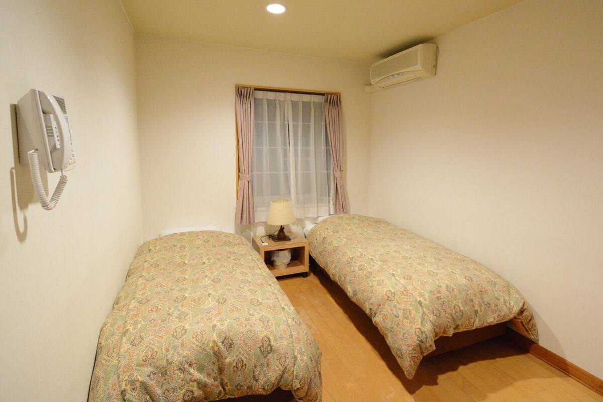 Ein kleines Zimmer mit zwei Einzelbetten, einem Nachttisch mit Lampe, einem Telefon und einer Klimaanlage.