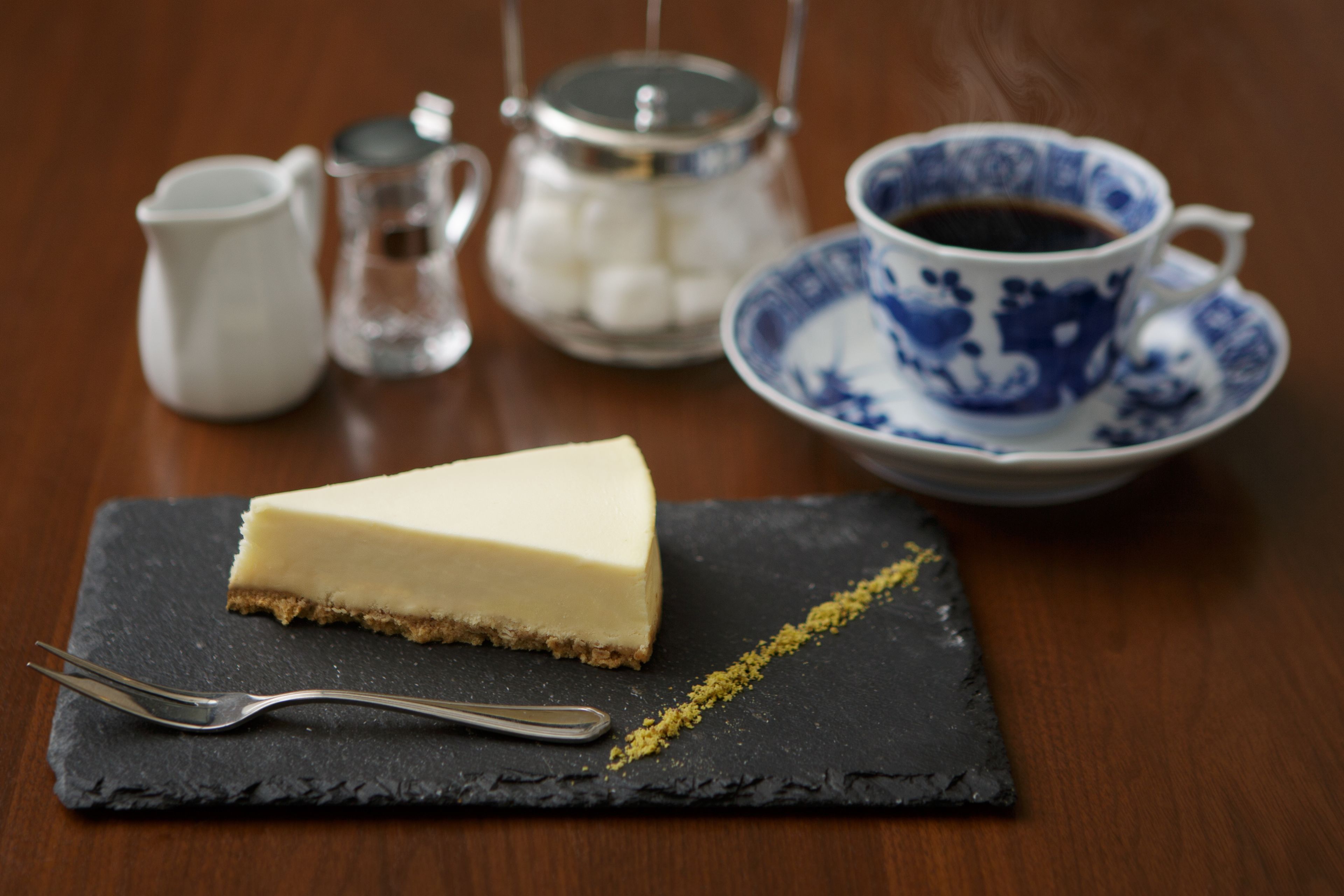 A slice of cheesecake on a slate plate with a fork, a cup of coffee, a teapot, and a sugar bowl on a wooden table.