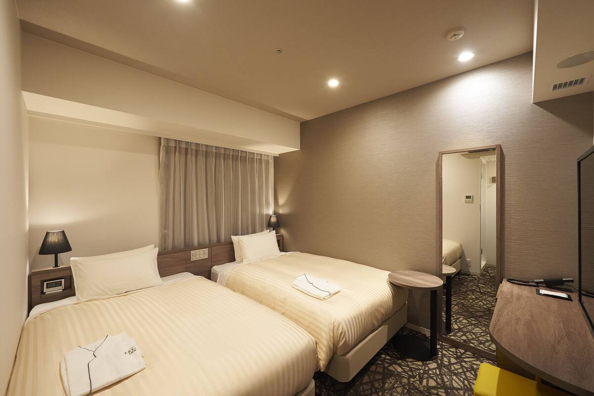 A hotel room with two beds, a bedside table with a lamp, a small round table, and a mirror.