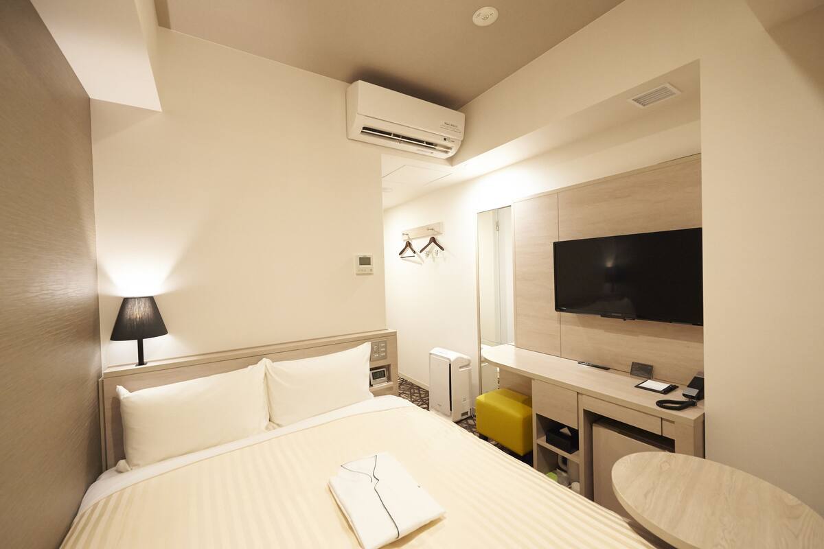 A hotel room with a bed, bedside lamp, air conditioning unit, TV, desk, and a mirror.