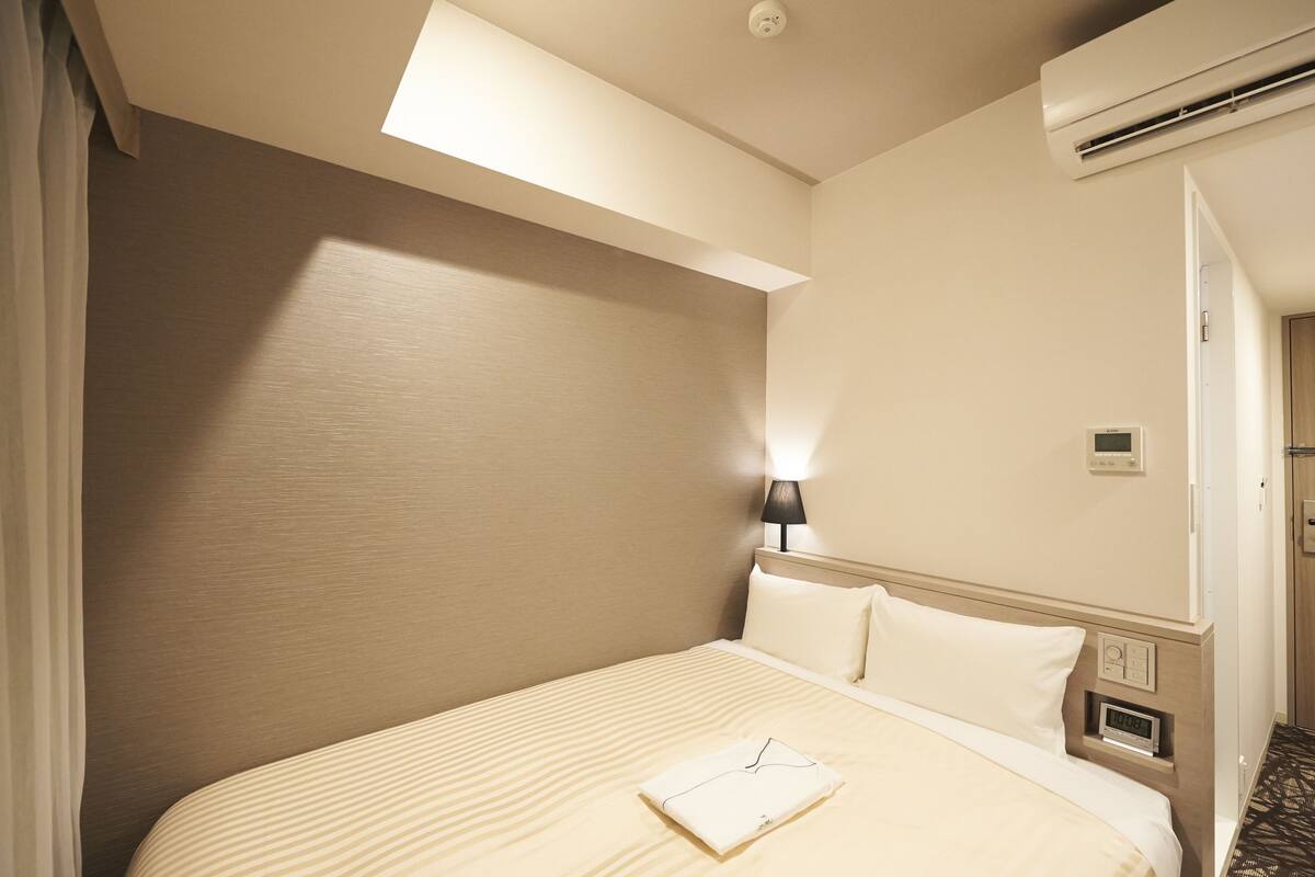 A hotel room with a bed, a nightstand, a lamp, and an air conditioning unit.
