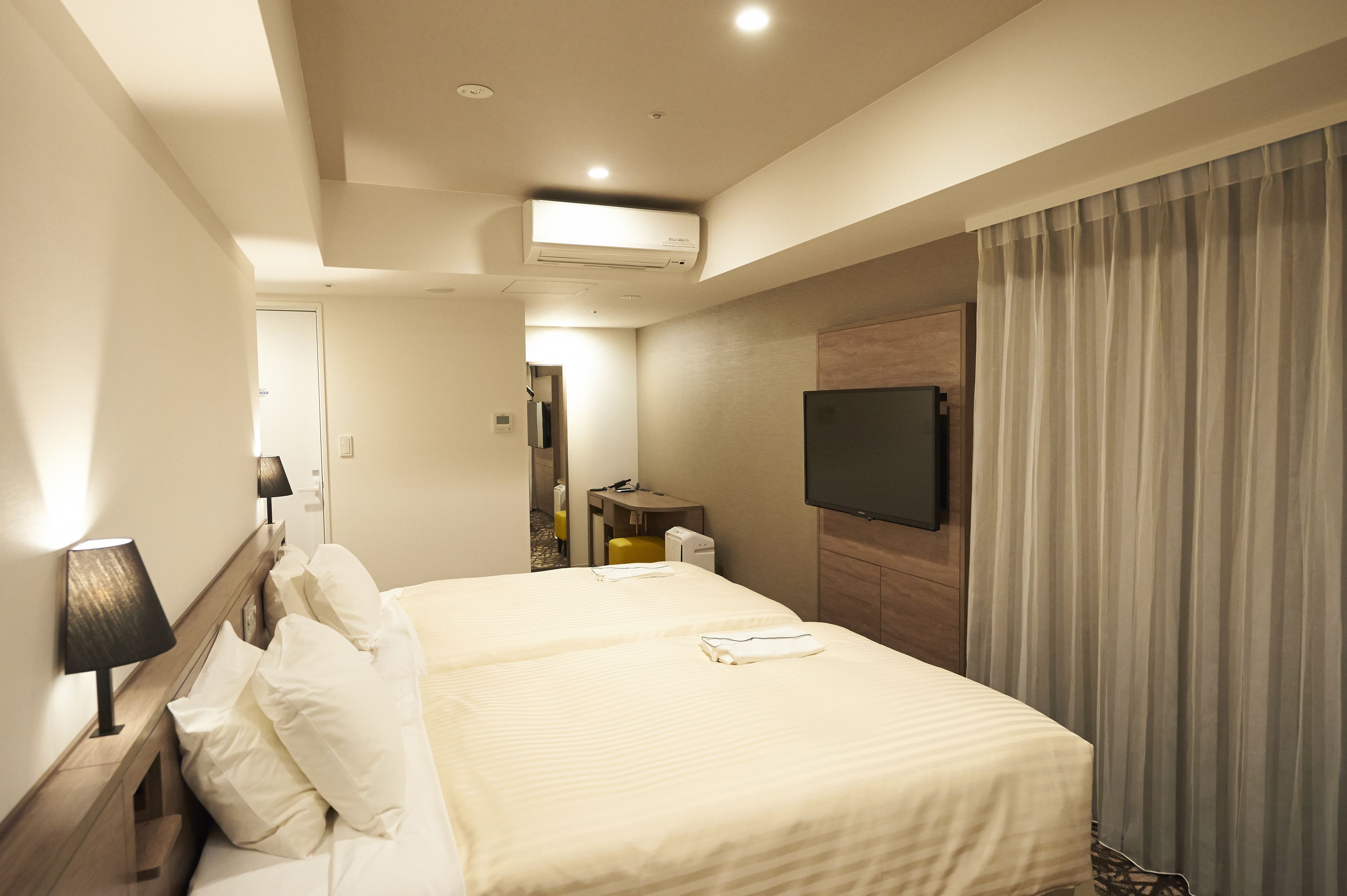 A hotel room with two beds, a flat-screen TV mounted on the wall, an air conditioning unit, a desk with a chair, and a curtain on the window.