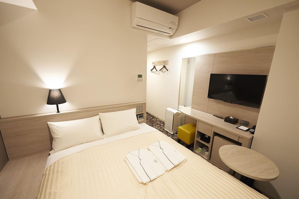 A hotel room with a bed, bedside lamp, air conditioning unit, TV, desk, chair, and a small round table.