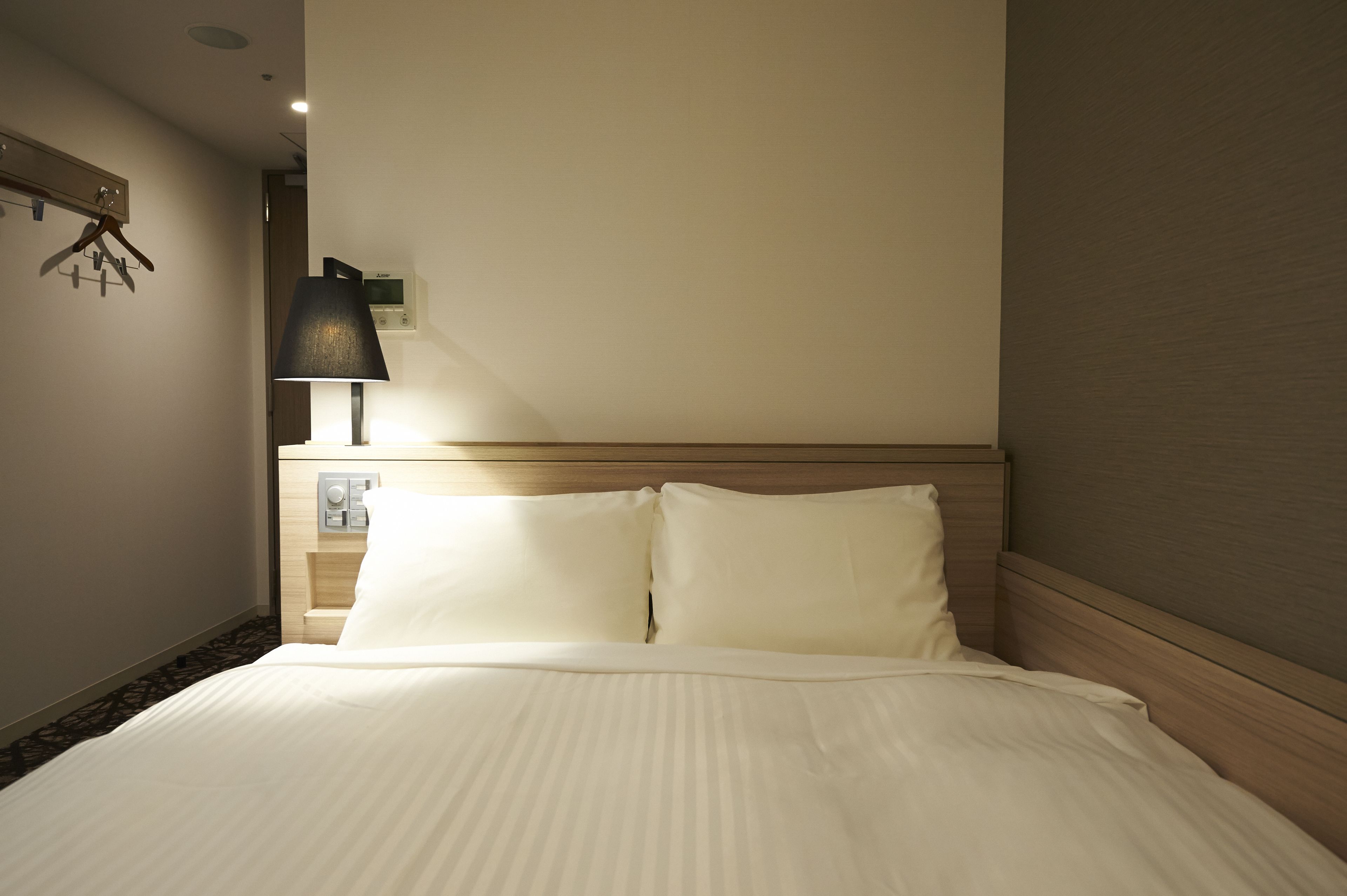 A hotel room with a neatly made bed, a bedside table with a lamp, and a wall-mounted heater.