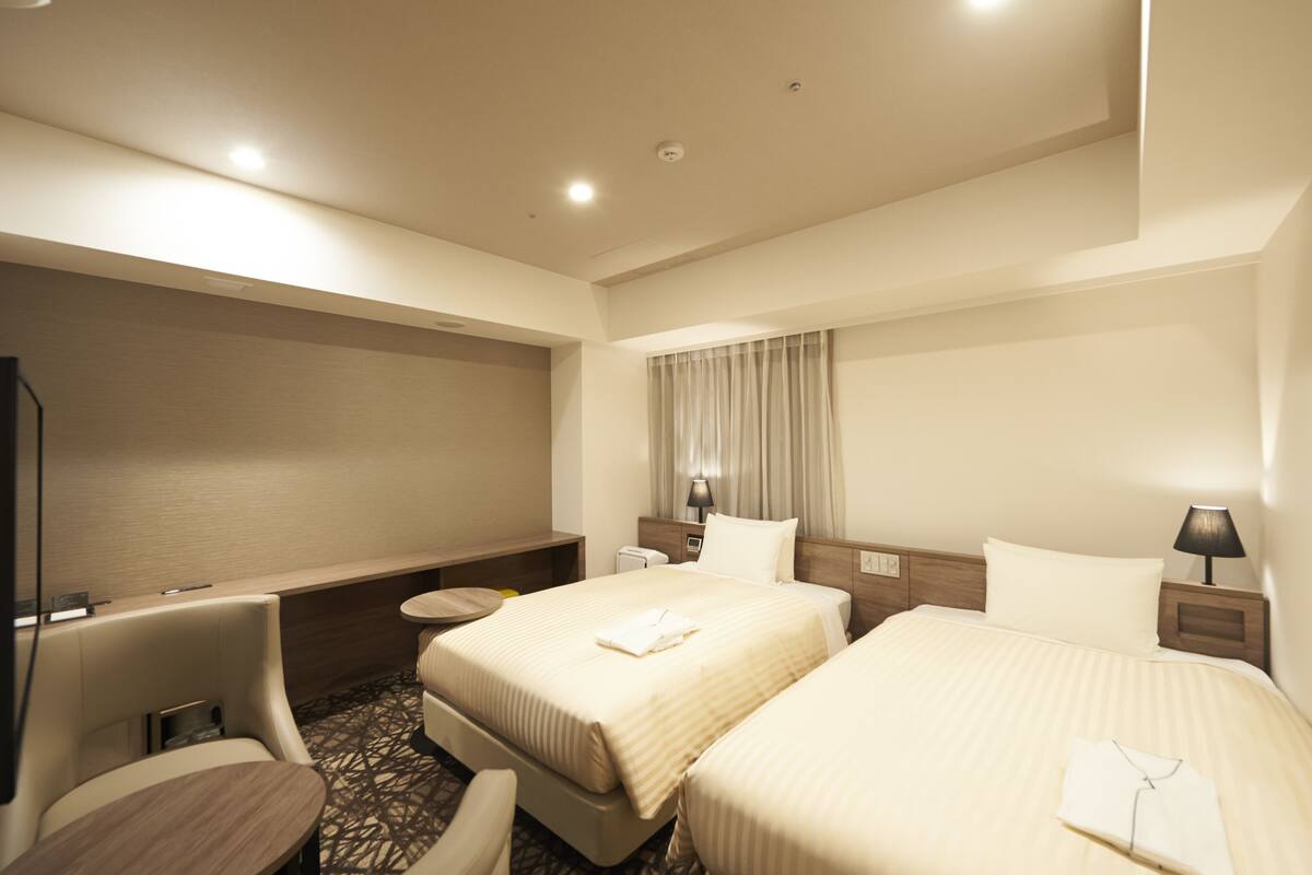 A hotel room with two beds, a desk, a chair, and a TV.