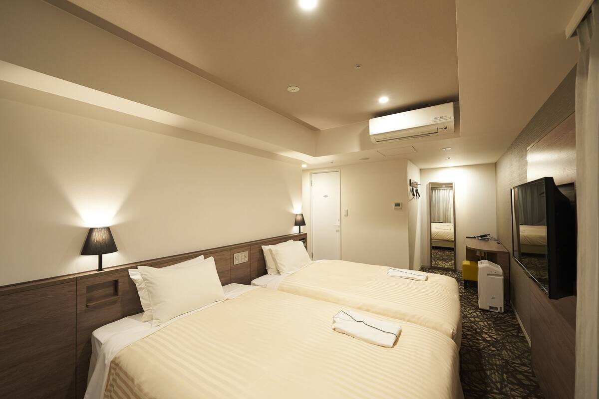 A hotel room with two beds, a desk, a chair, a TV, and an air conditioning unit.