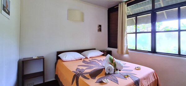 Uma cama bem arrumada com colcha florida, uma pequena mesa de cabeceira e uma janela com cortinas.