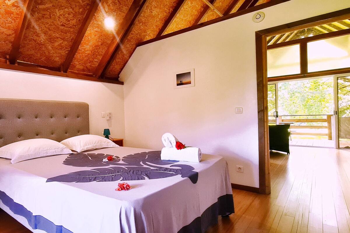 A bedroom with a large bed, a bedside table, a nightstand, a wall-mounted lamp, and a window with a view of greenery.