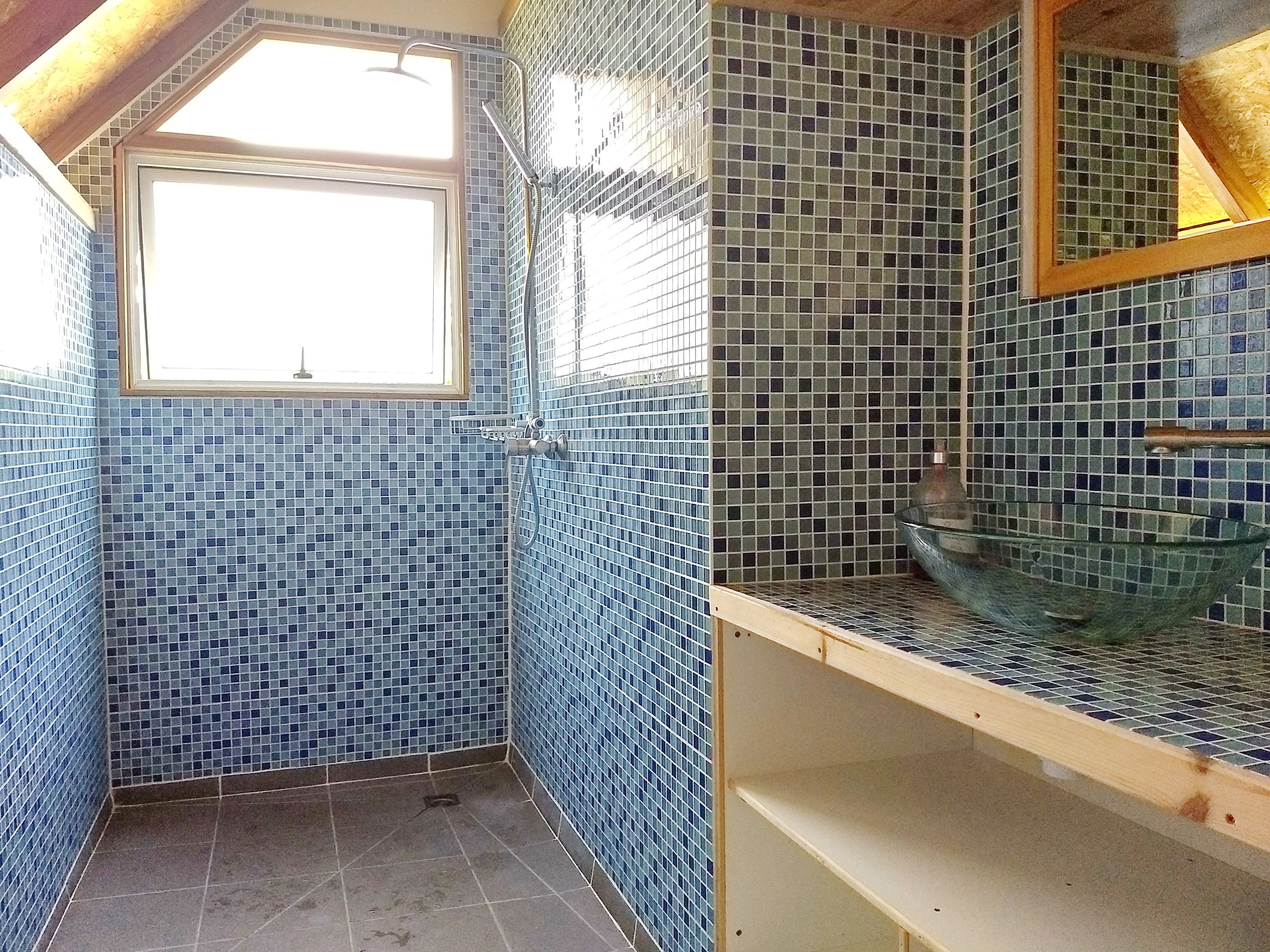 A bathroom with blue mosaic tiles, a window, a shower, and a glass sink.