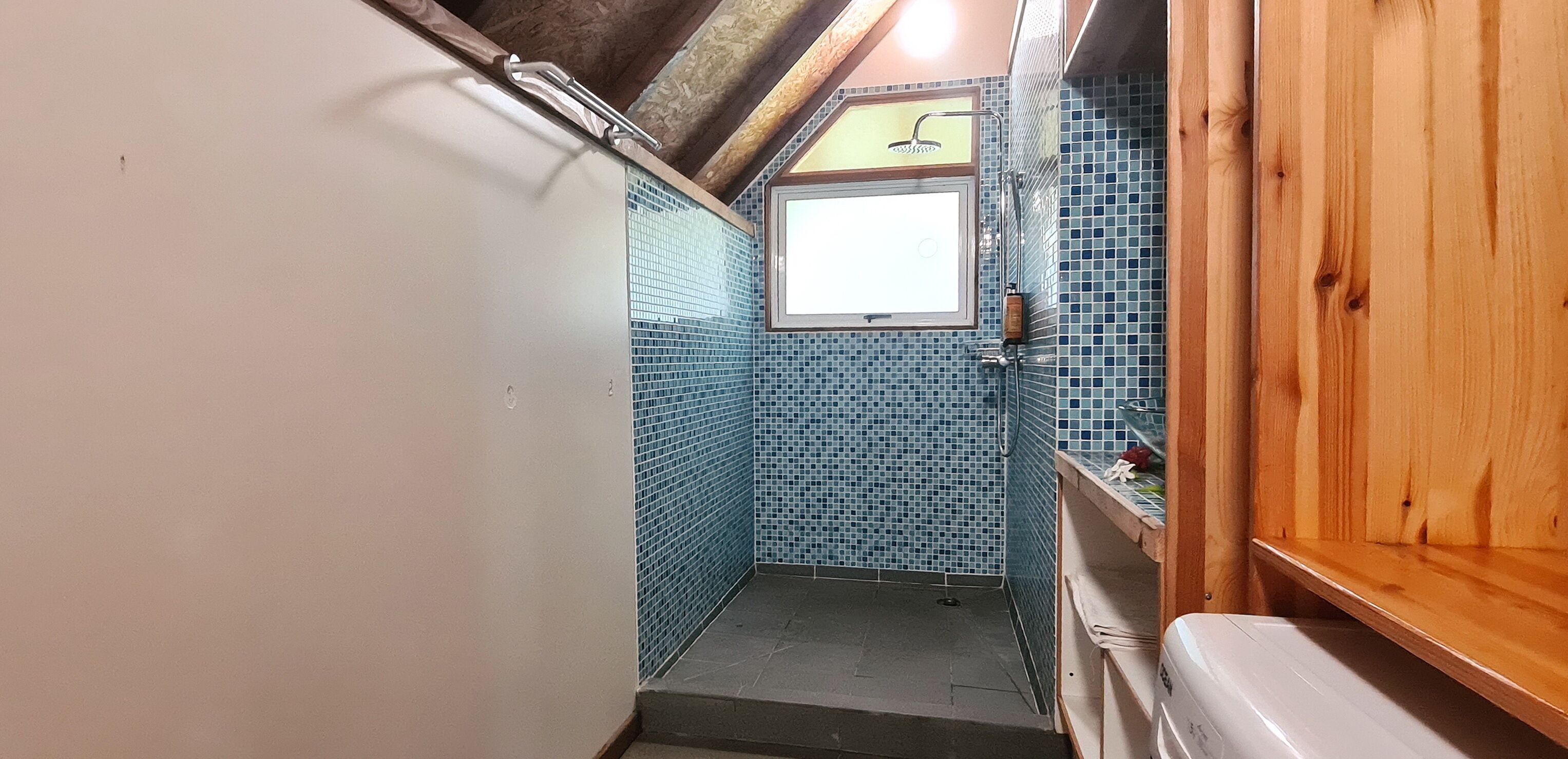 Et smalt badeværelse med blå mosaikfliser, et vindue og træhylder.