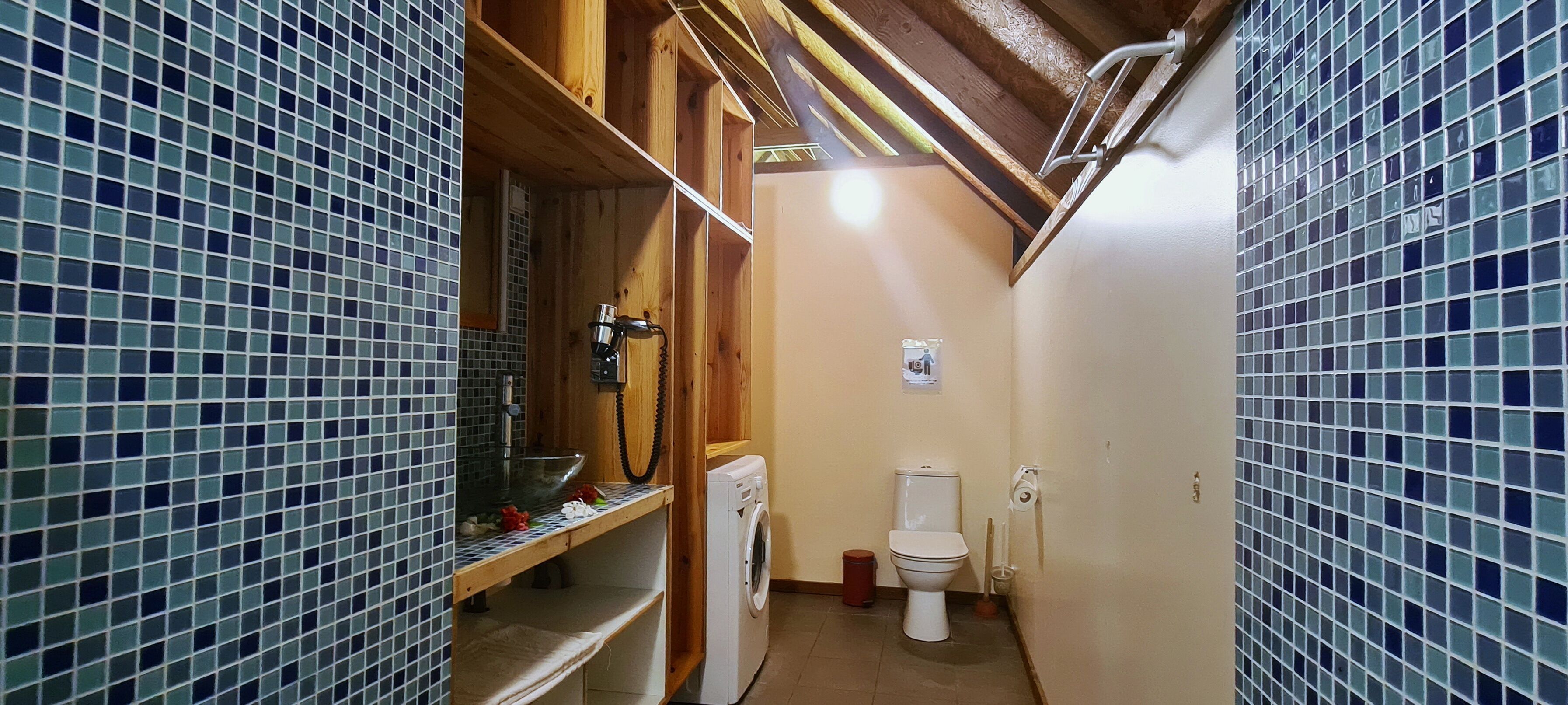 A compact bathroom with a wooden shelf, a washing machine, a toilet, and a tiled wall.