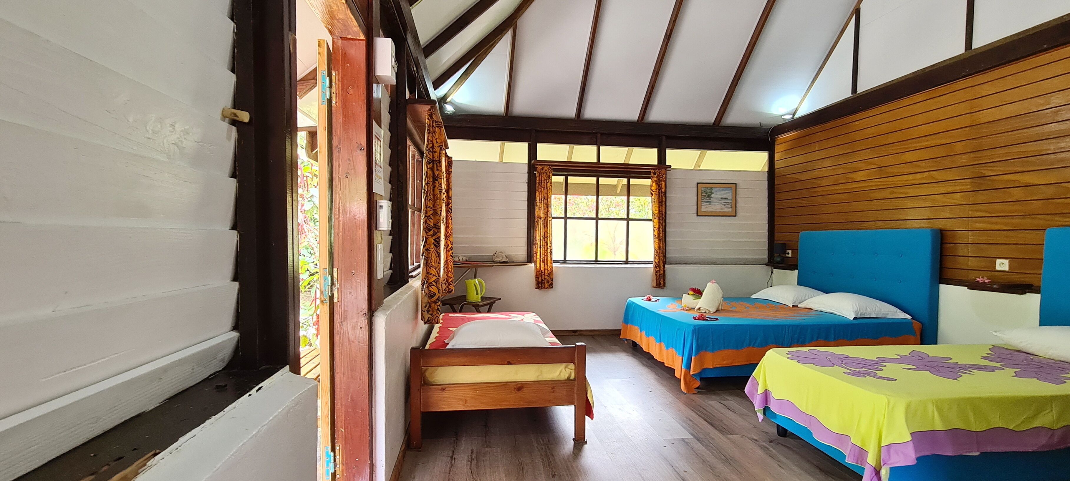 A room with two beds, wooden walls, a window with curtains, and a small table with a kettle.