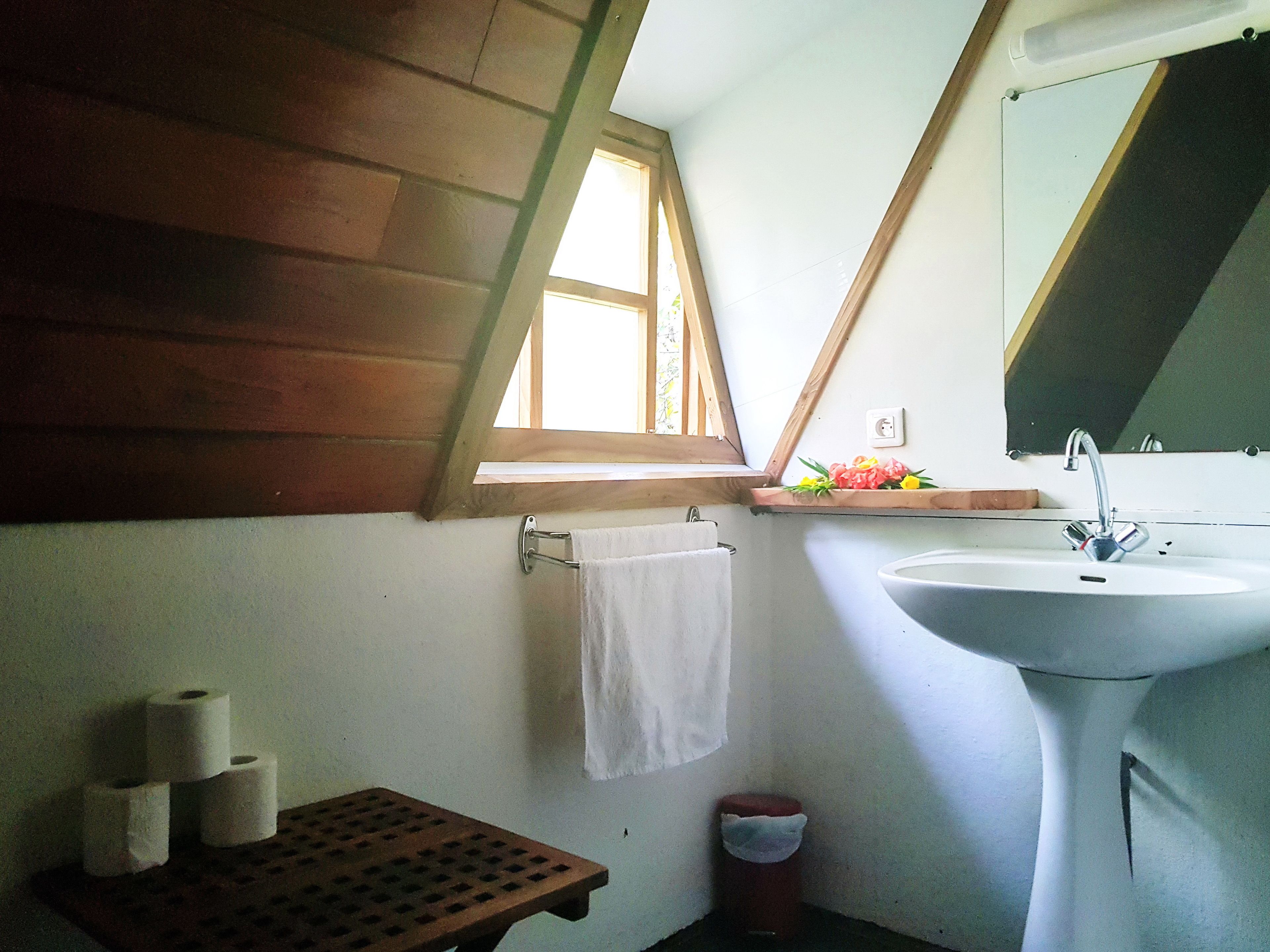 Un baño con techo de madera, una ventana, un lavamanos y una pequeña estantería con flores.
