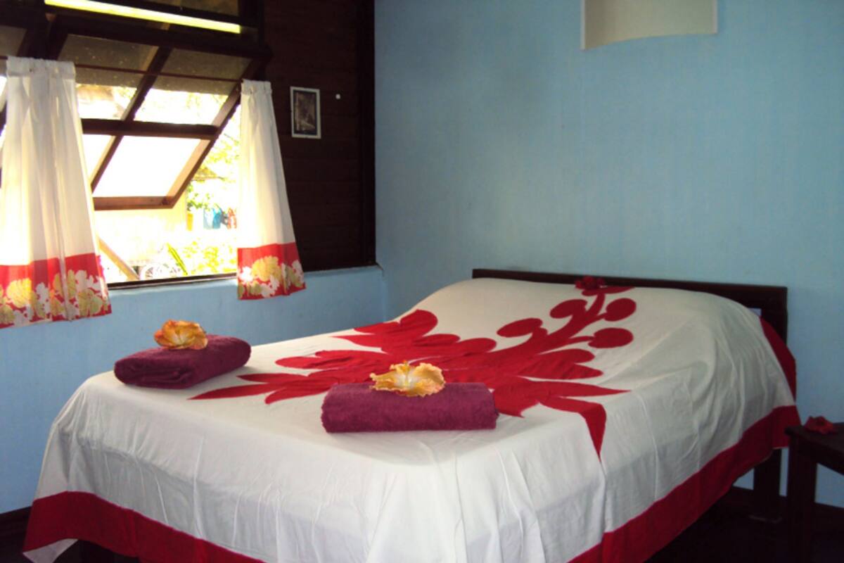 A neatly made bed with a white and red floral bedspread, two purple towels, and a window with curtains.