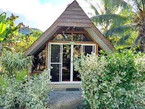 A small wooden structure with a triangular roof and a sliding glass door, surrounded by lush greenery and palm trees.
