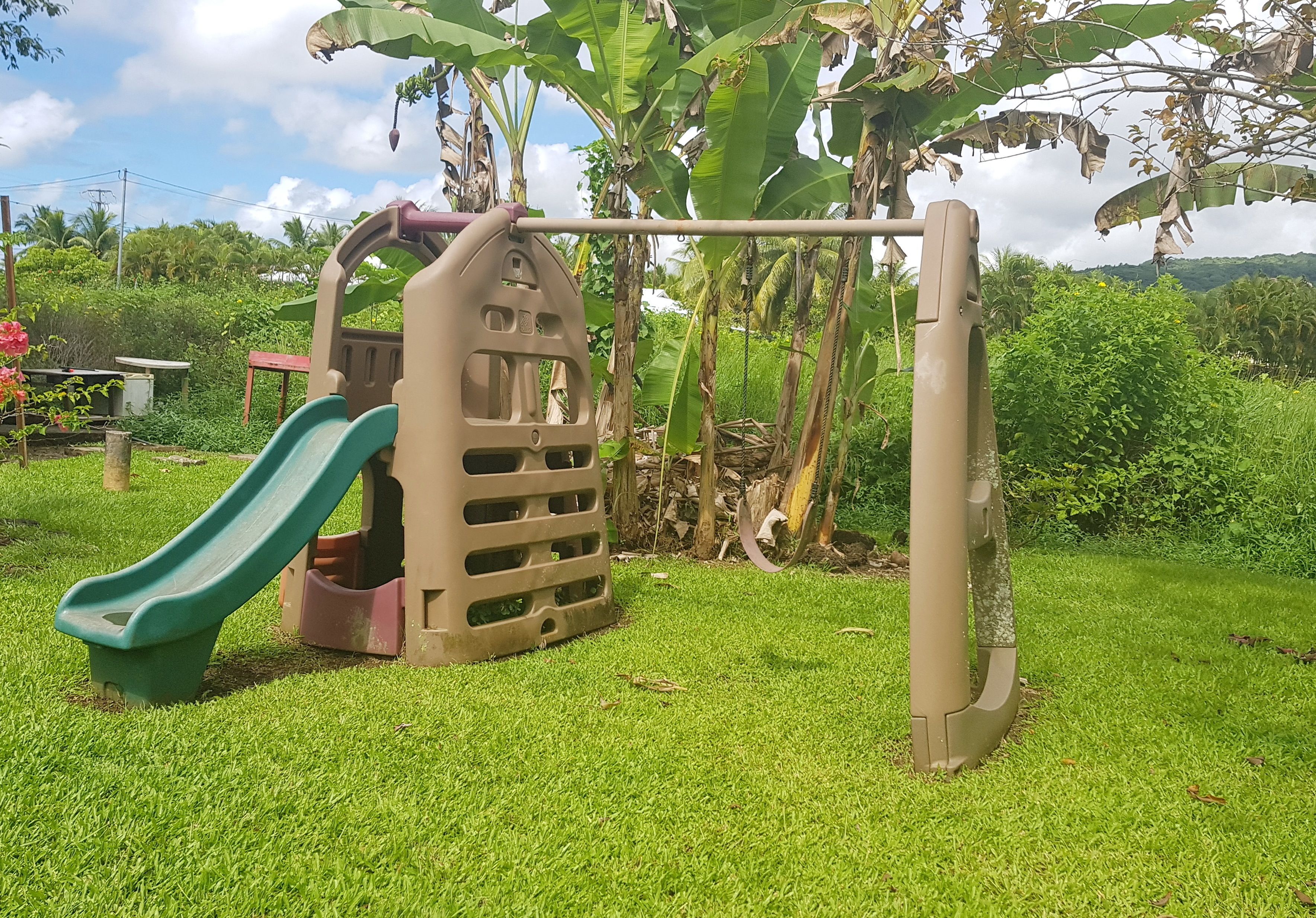 Une aire de jeux avec un toboggan et une structure d’escalade, située dans un espace verdoyant avec des arbres et un ciel dégagé.