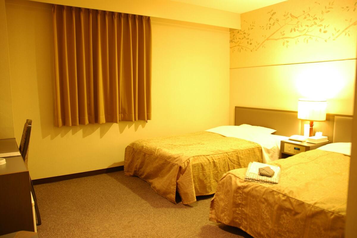 A hotel room with two beds, a desk, a chair, a lamp, and a wall with a branch design.