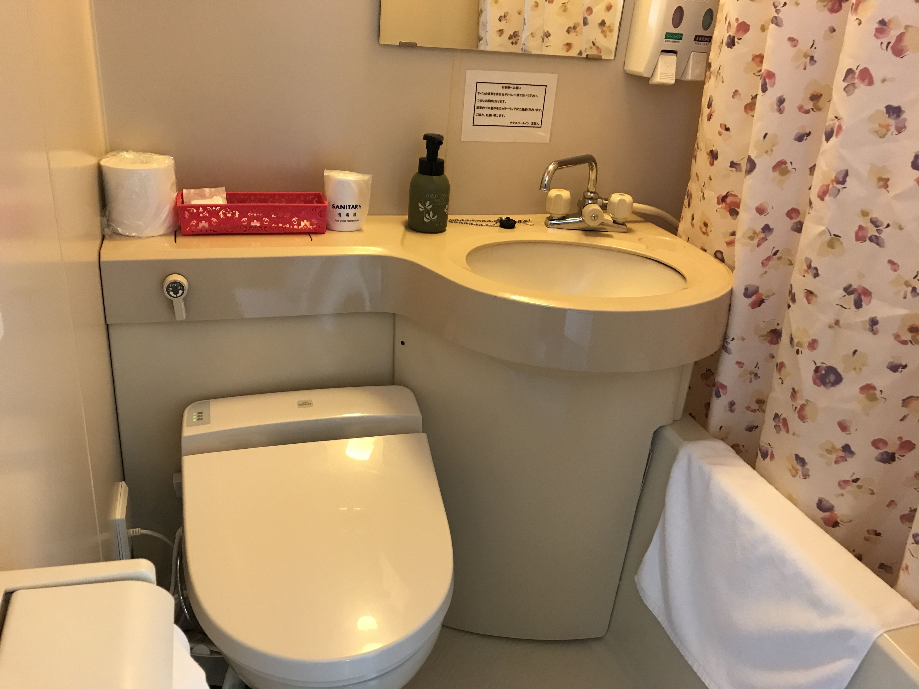 Ein Badezimmer mit Toilette, Waschbecken und Duschvorhang. Auf der Ablage des Waschbeckens stehen Toilettenartikel.