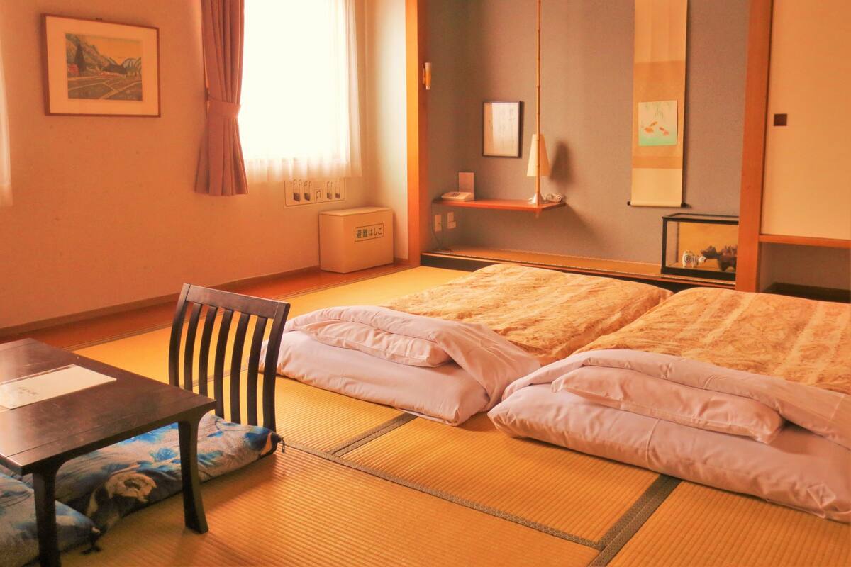 A traditional Japanese room with tatami flooring, a low table, a chair, and a bed with pillows.