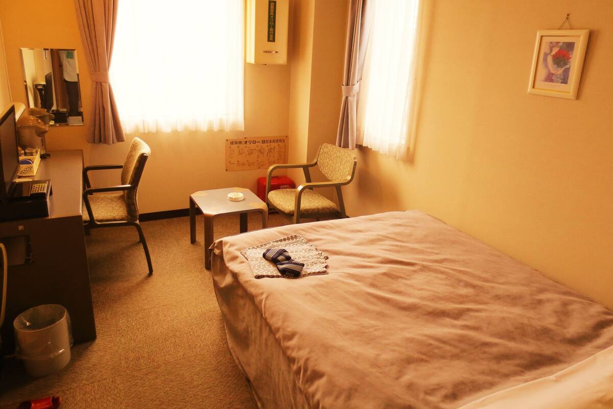 A hotel room with a bed, two chairs, a desk with a telephone, a small table, and a picture on the wall.