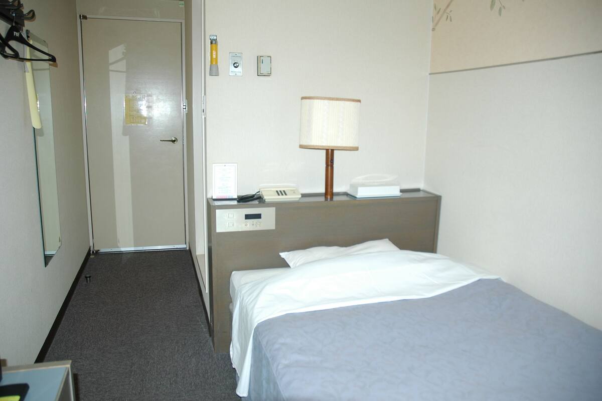 A hotel room with a bed, a desk with a lamp, a telephone, and a door.