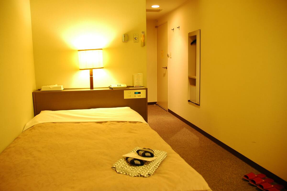 A hotel room with a bed, a bedside table with a lamp, a mirror, and a door leading to another room.