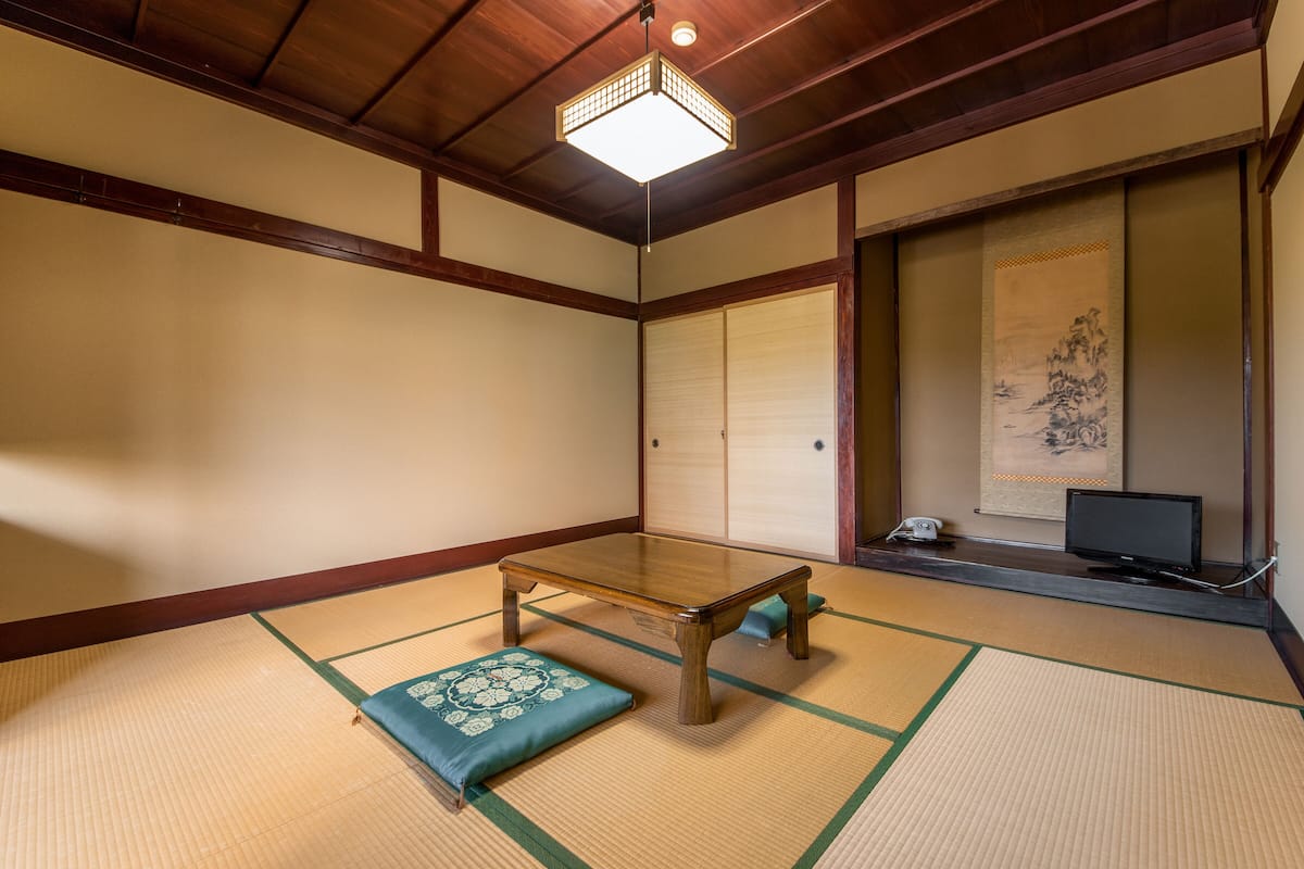 Ein traditioneller japanischer Raum mit Tatami-Bodenbelag, einem niedrigen Holztisch, Kissen, einem Fernseher und einer Schiebetür.