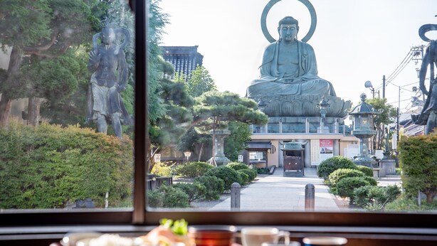Vista de una gran estatua de Buda desde el interior de un edificio, con una ventana que da a una calle bordeada de árboles y otras estatuas.