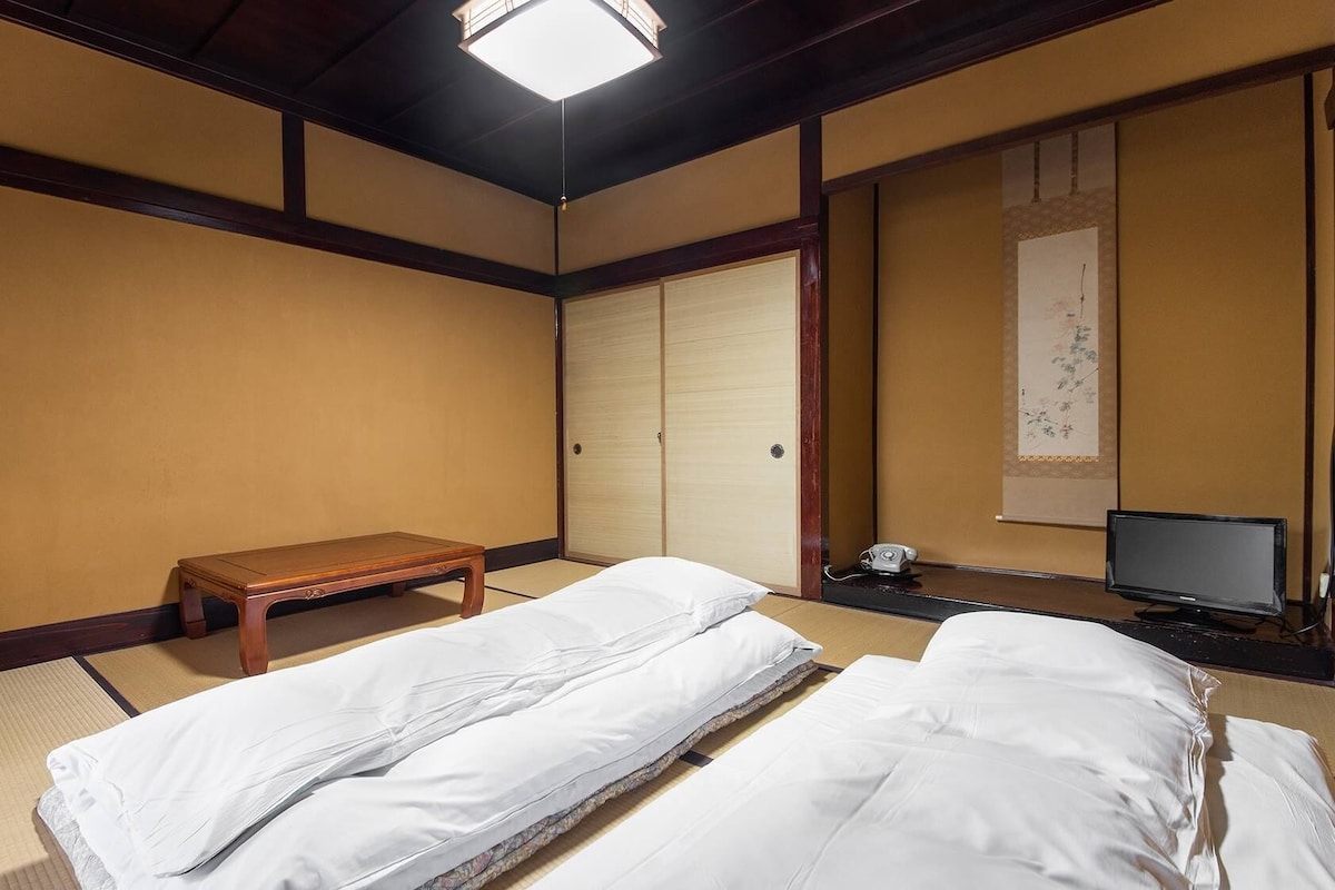 A traditional Japanese room with tatami flooring, a futon bed, a wooden bench, a sliding door, a desk with a telephone, and a television.