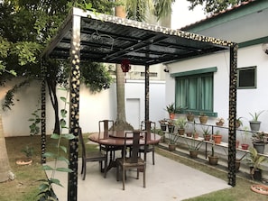 Un patio cubierto con una mesa redonda y sillas, rodeado de plantas en macetas y una pared blanca con marco de ventana verde.