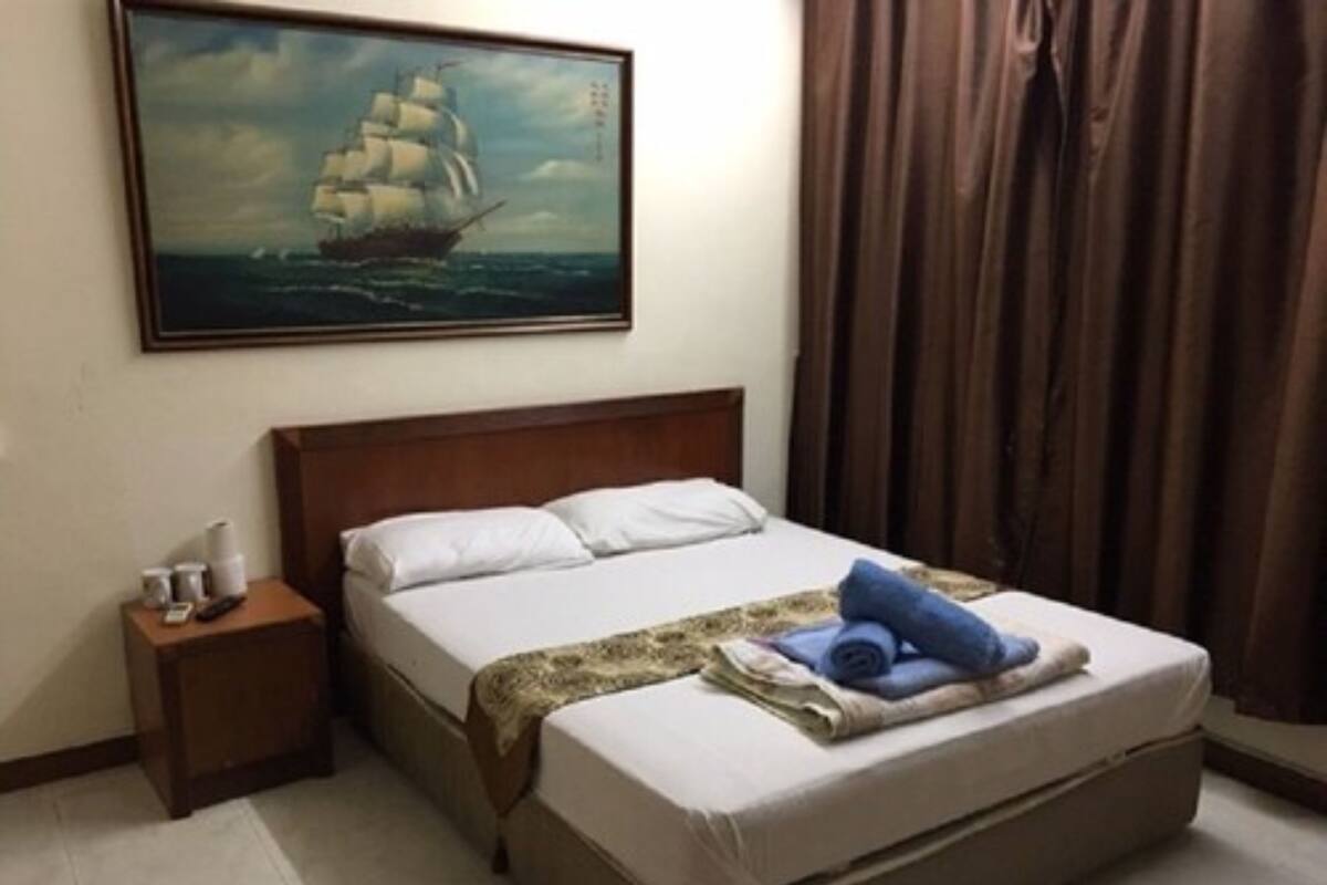 Een hotelkamer met een groot bed, een nachtkastje, een schilderij van een schip aan de muur en een raam met gordijnen.
