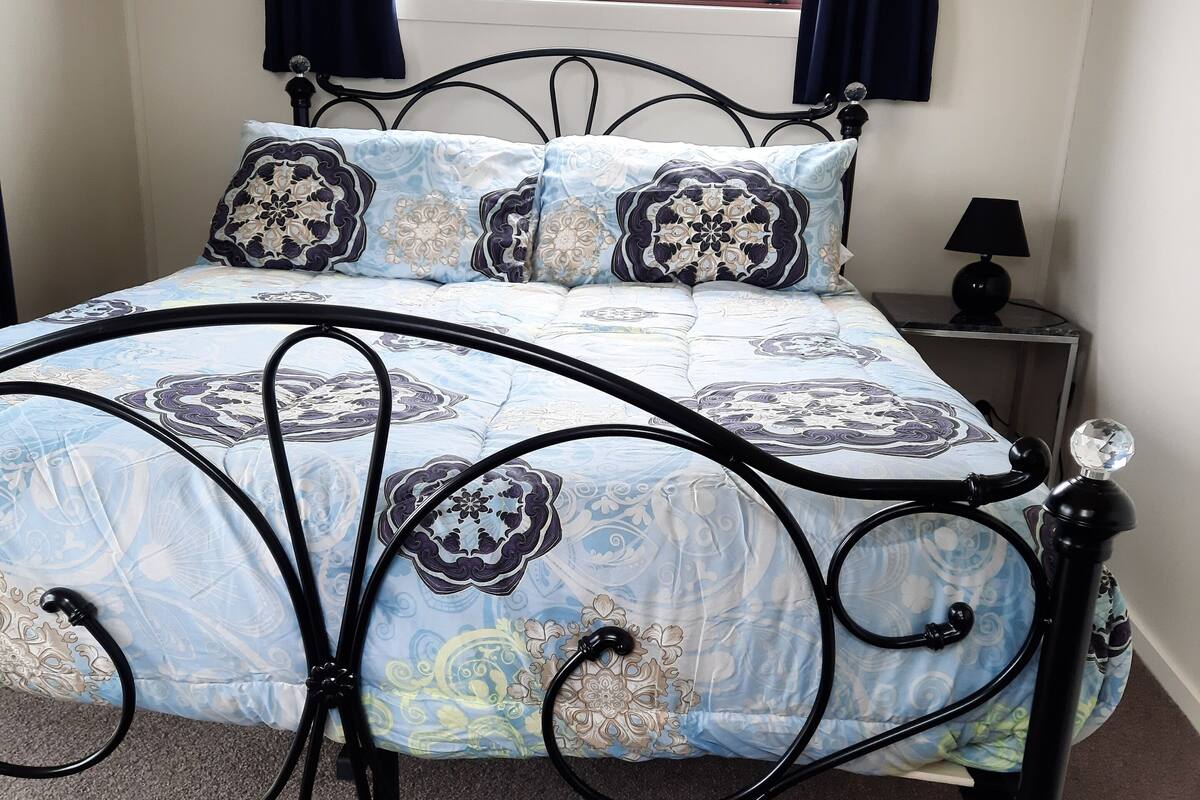 Ein Bett mit gemustertem Überwurf und passenden Kissen, einem schwarzen Metallgestell, einem Nachttisch mit Lampe und einem Fenster mit Vorhängen.