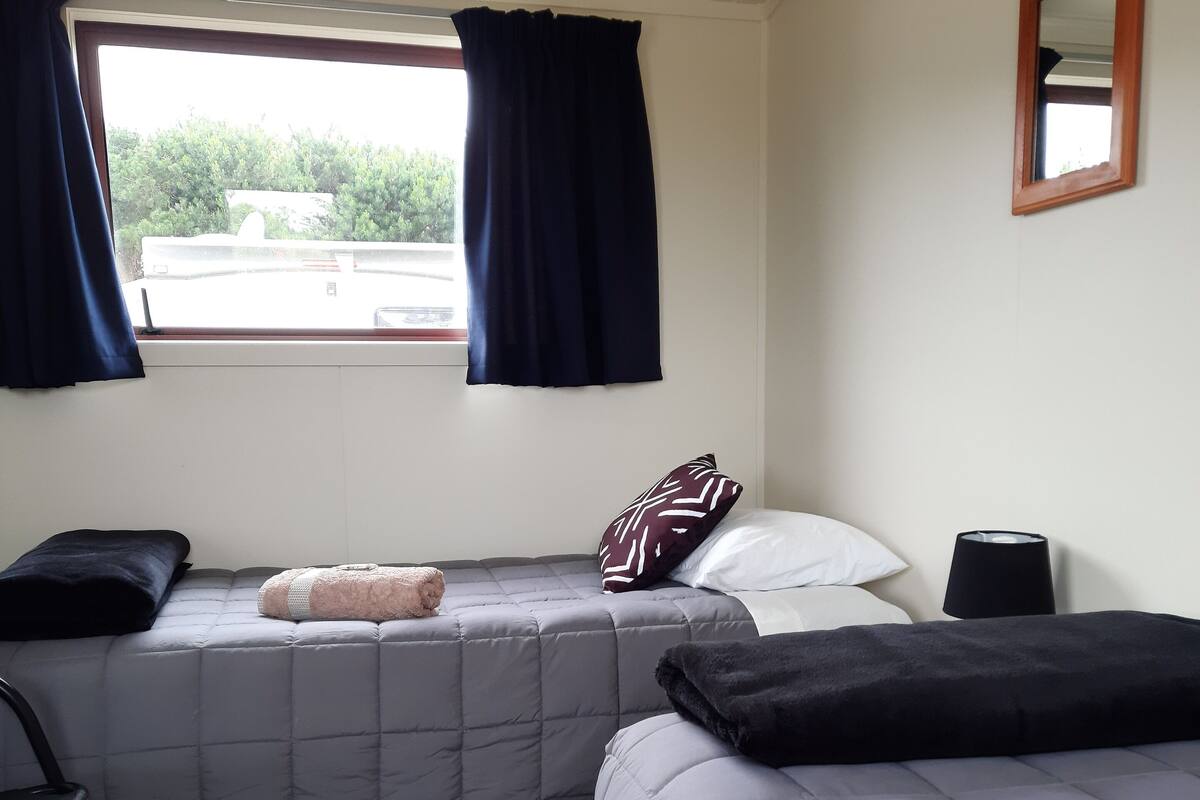 Ein Schlafzimmer mit zwei Betten, einem Fenster mit Vorhängen, einem Nachttisch und einem Sessel.