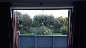 On aperçoit, à travers une porte-fenêtre coulissante, un jardin avec une terrasse en bois, une clôture en métal et divers arbustes et arbres.